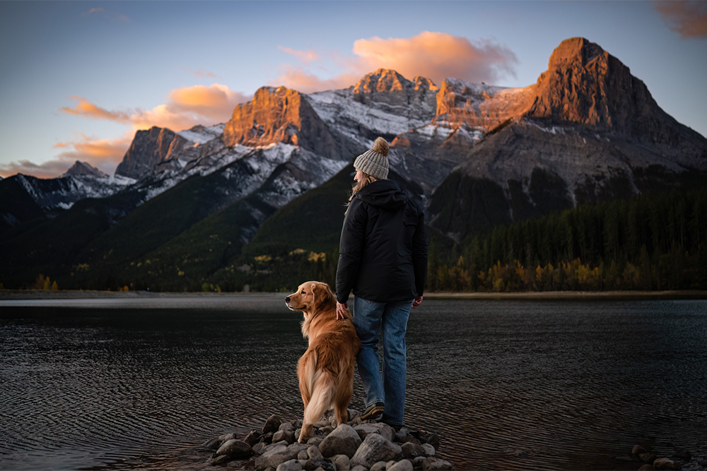 A woman and a golden retriever stand on the rocky shore of a lake or river facing tall mountains.