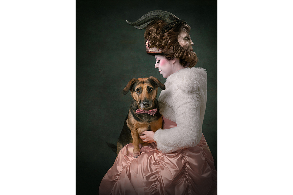A woman wearing a pink and white gown and a mask on the back of her head sits with a brown and black dog on her lap.