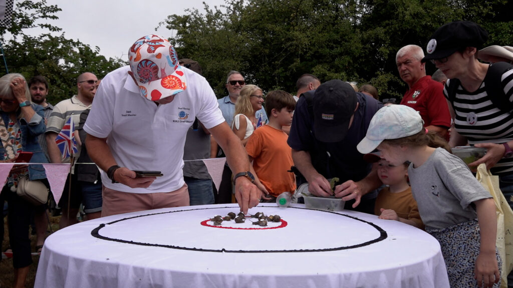 A man positions a group of snails within concentric circles on a table as a child watches and a crowd gathers in the background.
