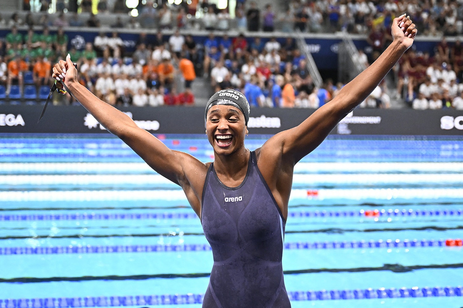 Sara Curtis smiles and has her arms raised as she stands in front of a pool in her swimsuit and cap.