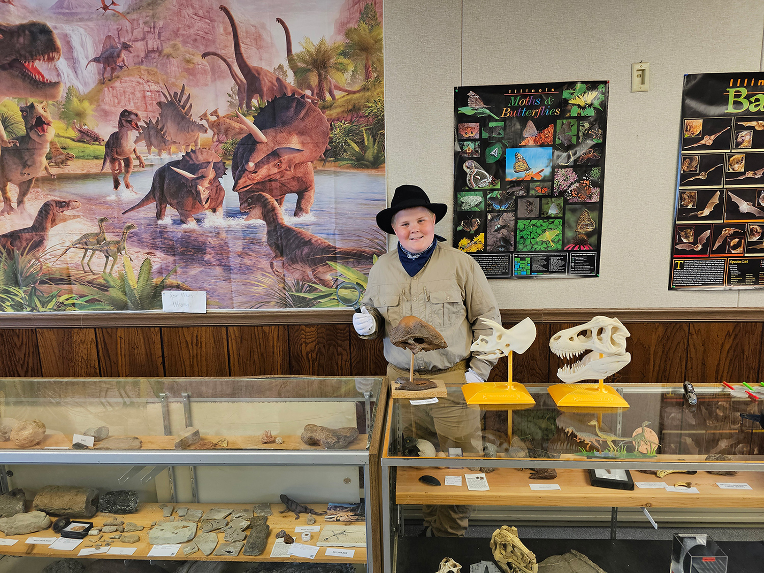 Anderson Taylor stands behind display cases of fossils and other objects with posters of dinosaurs, moths, and butterflies in the background.