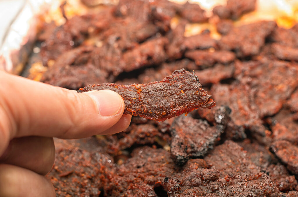 A hand is holding a piece of jerky over a pile of jerky.