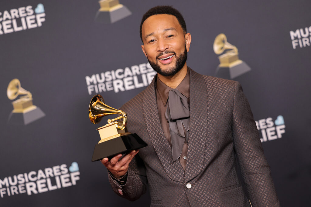 John Legend poses in front of a background reading “MusicCares Fire Relief” while holding a Grammy award.