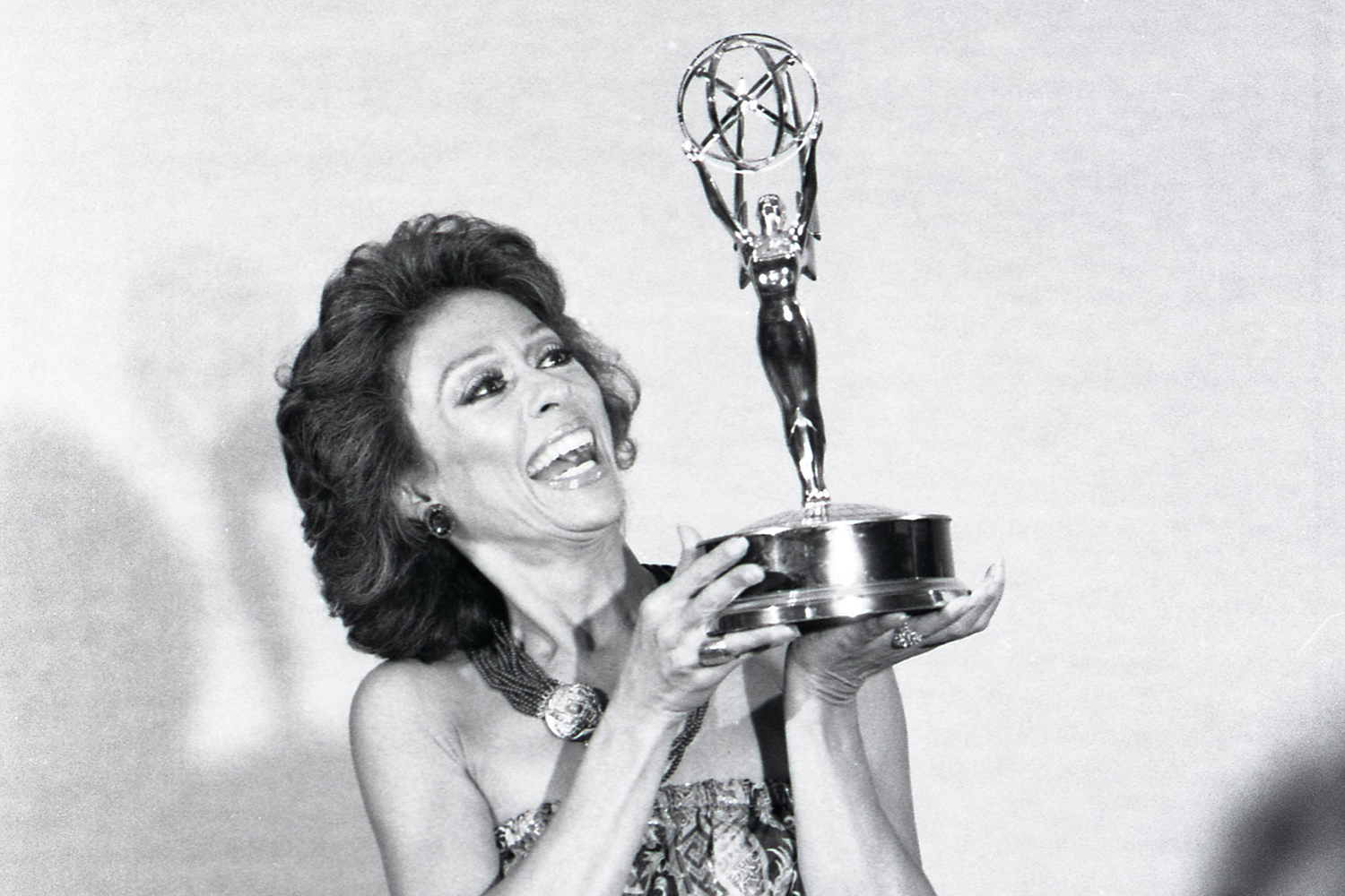 Rita Morena holds up an Emmy Award and smiles.