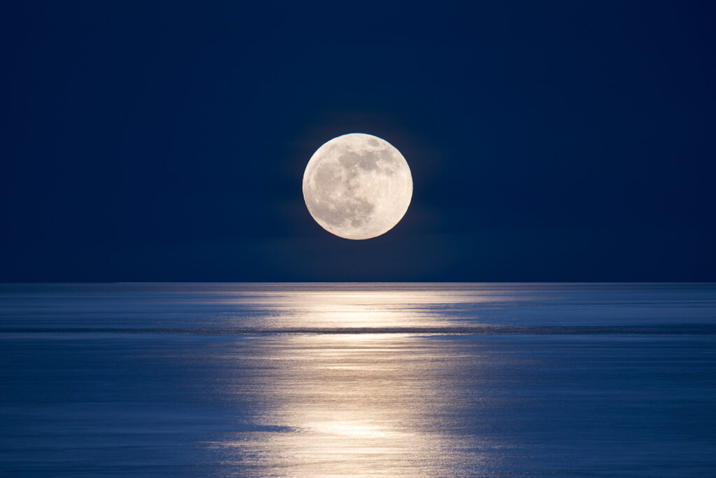 A full Moon rises over a body of water.
