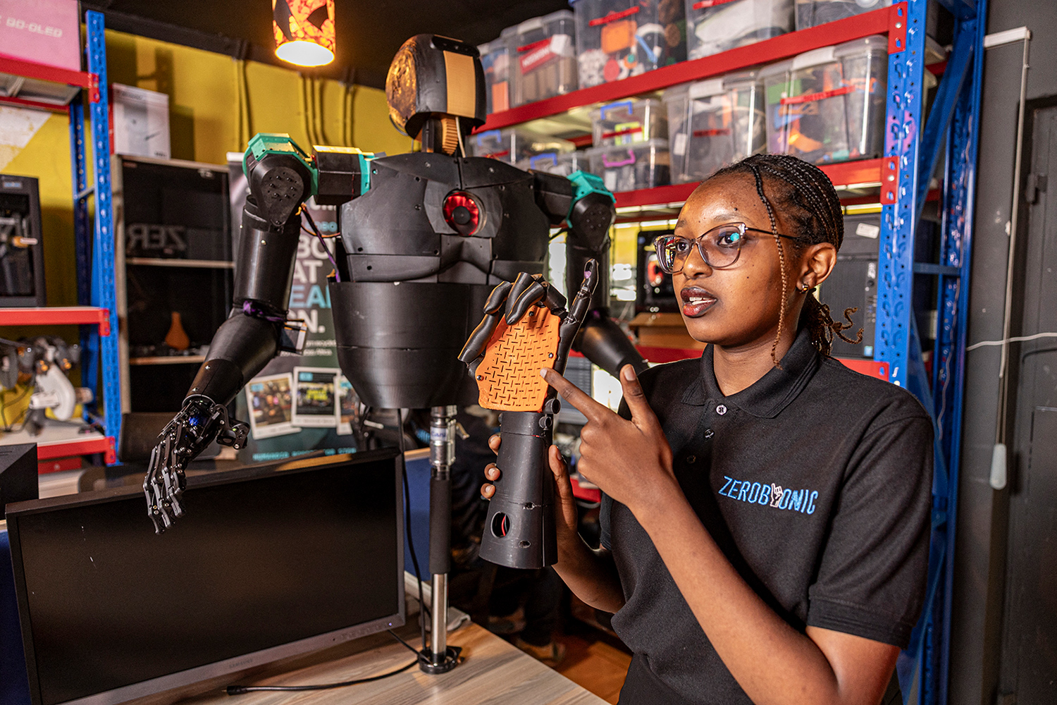 Norah Kimathi stands at a table and holds a robotic arm. Part of a humanoid robot is perched on the table.