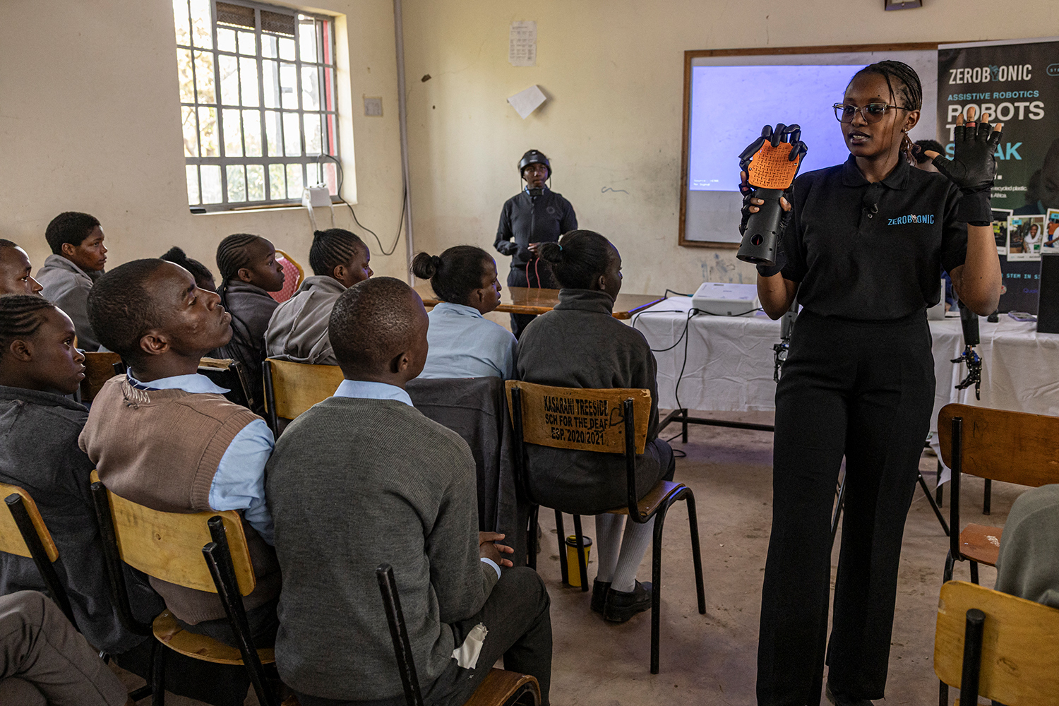 Norah Kimathi stands among seated students in a classroom and holds up a robotic arm.