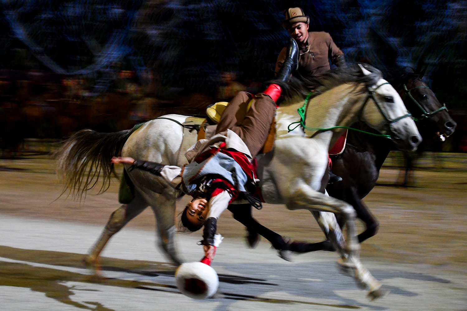 A horseback rider holds onto another rider who hangs over her moving horse on her back while holding an object.