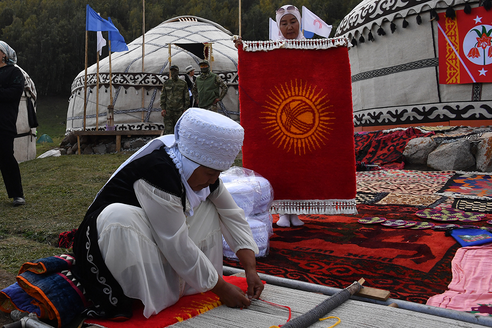 A person weaves on a loom on the ground as a woman holds up a woven rug.