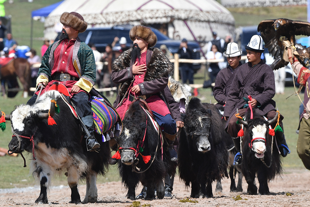 Four people in traditional clothing ride yaks with a crowd in the background.