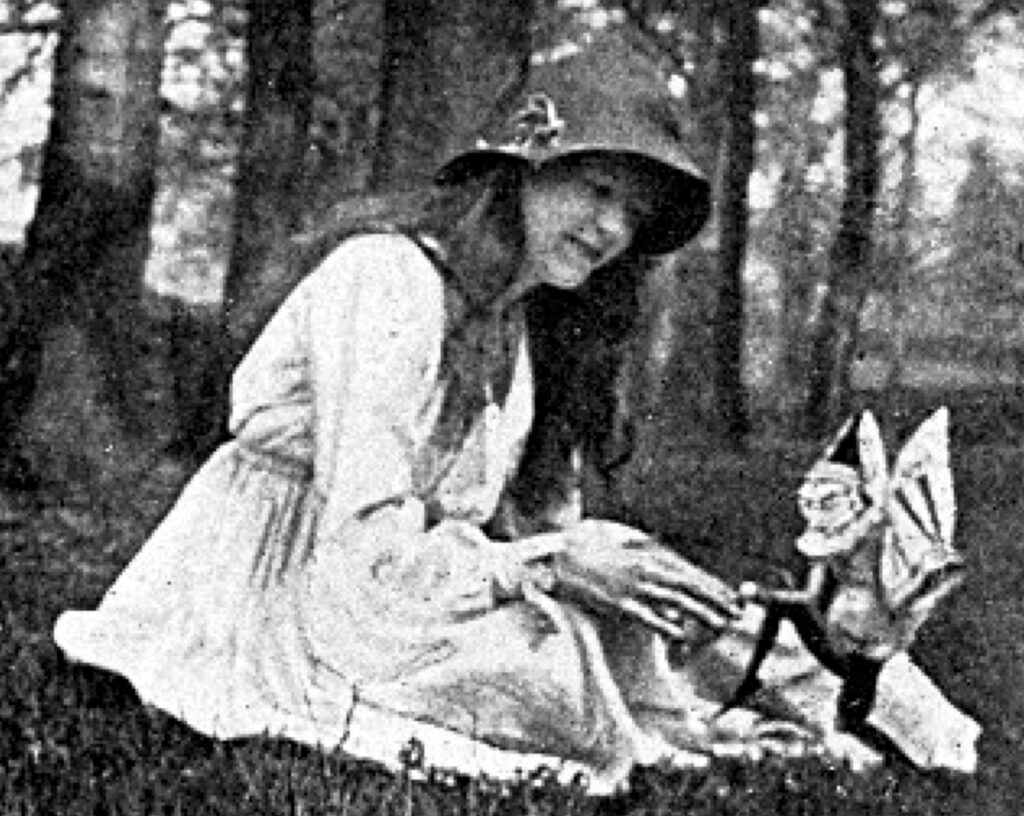 A woman in early 20th century clothing sits on grass and appears to be shaking hands with a tiny fairy creature.