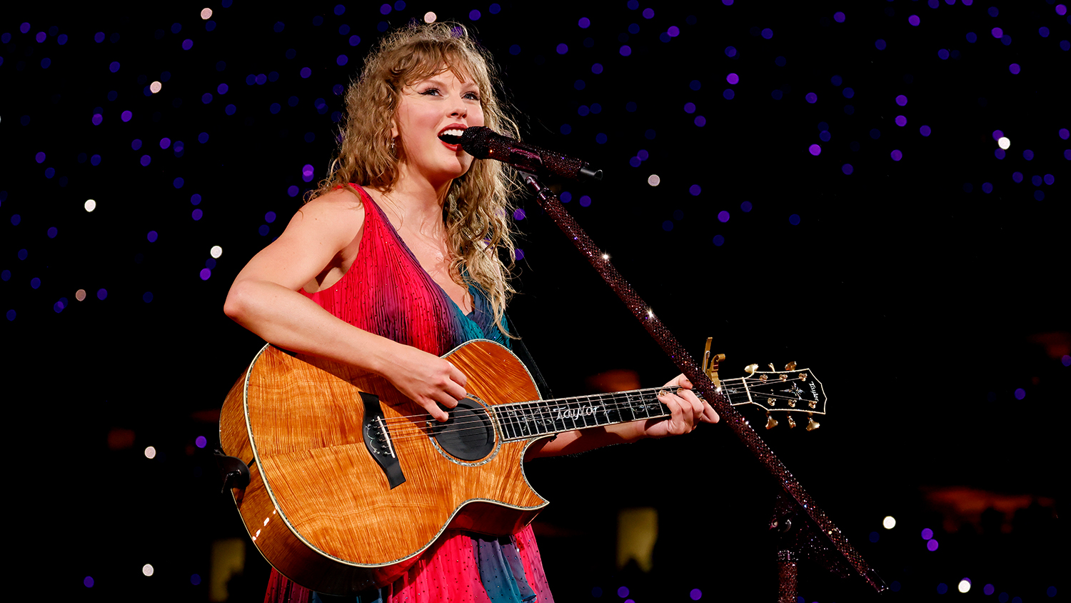 Taylor Swift stands on stage and smiles as she plays a guitar.