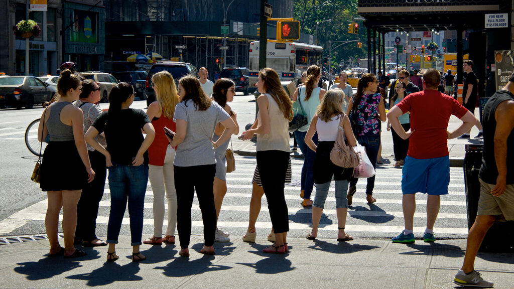 A large number of people are walking across a city street even though there is a red hand in the walk signal, indicating that pedestrians should not cross.