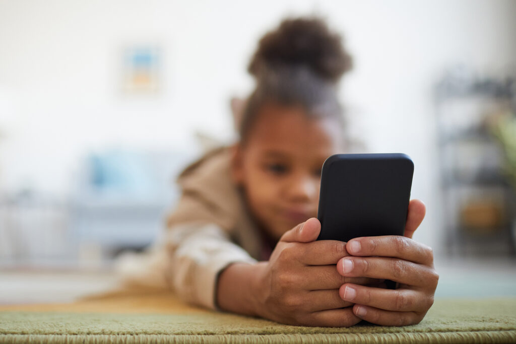 A young child lies on her stomach on the floor and looks at a smartphone.