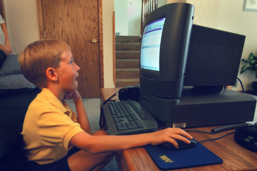 A young child sits at a desk and looks at a 1990s computer with a large monitor.