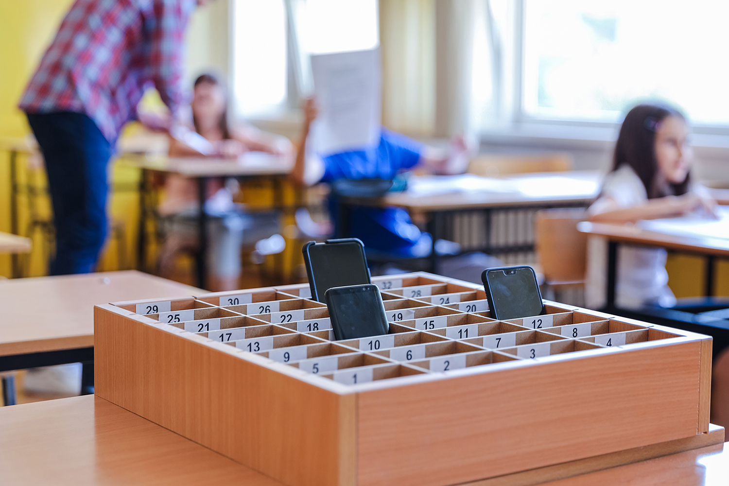 A wood box with numbered slots, some containing phones, sits in a classroom with elementary age students in the background.