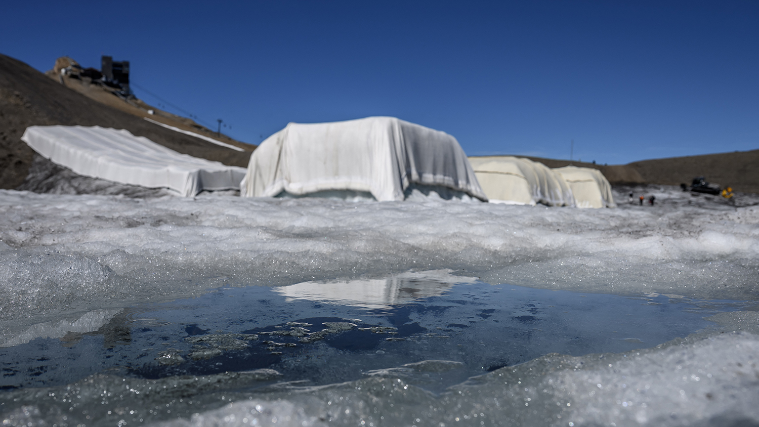 Ice melts in the sun in front of four large pieces of ice that are covered by tarps or wool.