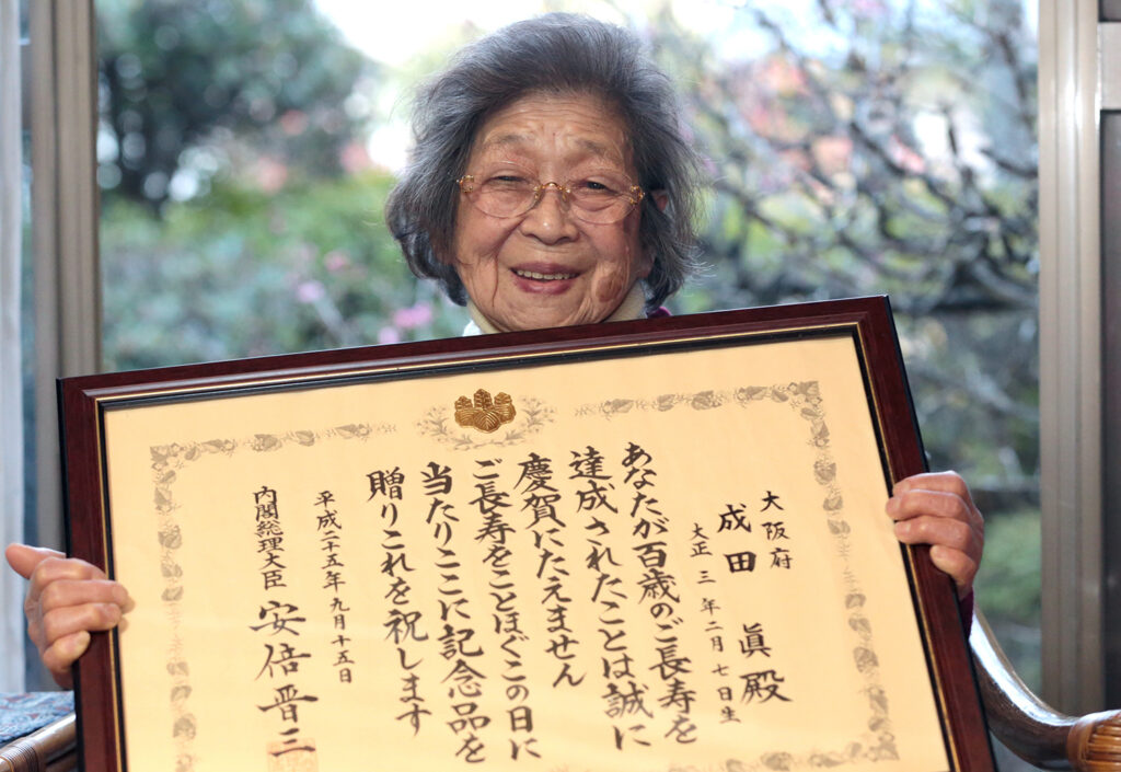 An older woman smiles and holds up a framed certificate that is written in Japanese.