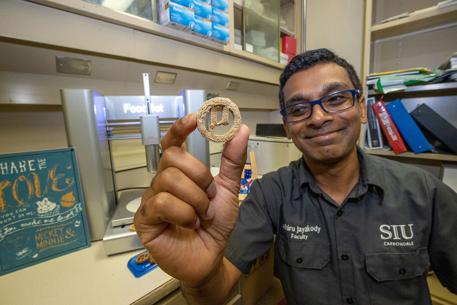 Lahiru Jayakody sits in a room with some books and equipment and holds up a round cookie.