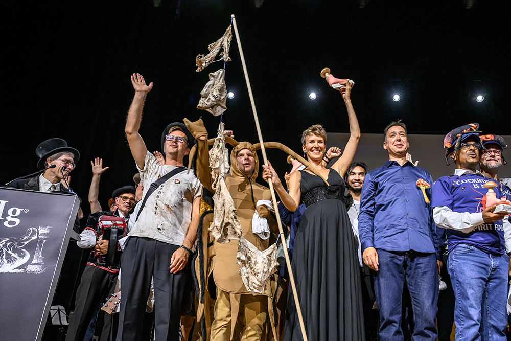 A group of people smile and wave on a stage. One of them is holding a line to which underwear is attached, and several are wearing cockroach-themed clothing.