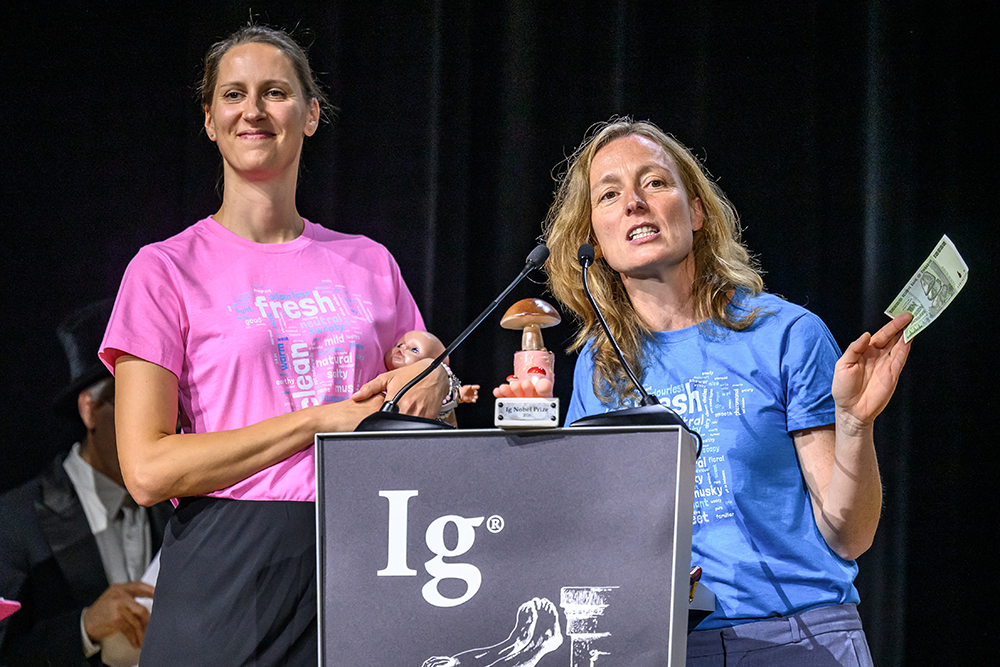 Two women stand at the Ig Nobel podium, one holding a baby doll and the other holding a trophy that looks like a mushroom.
