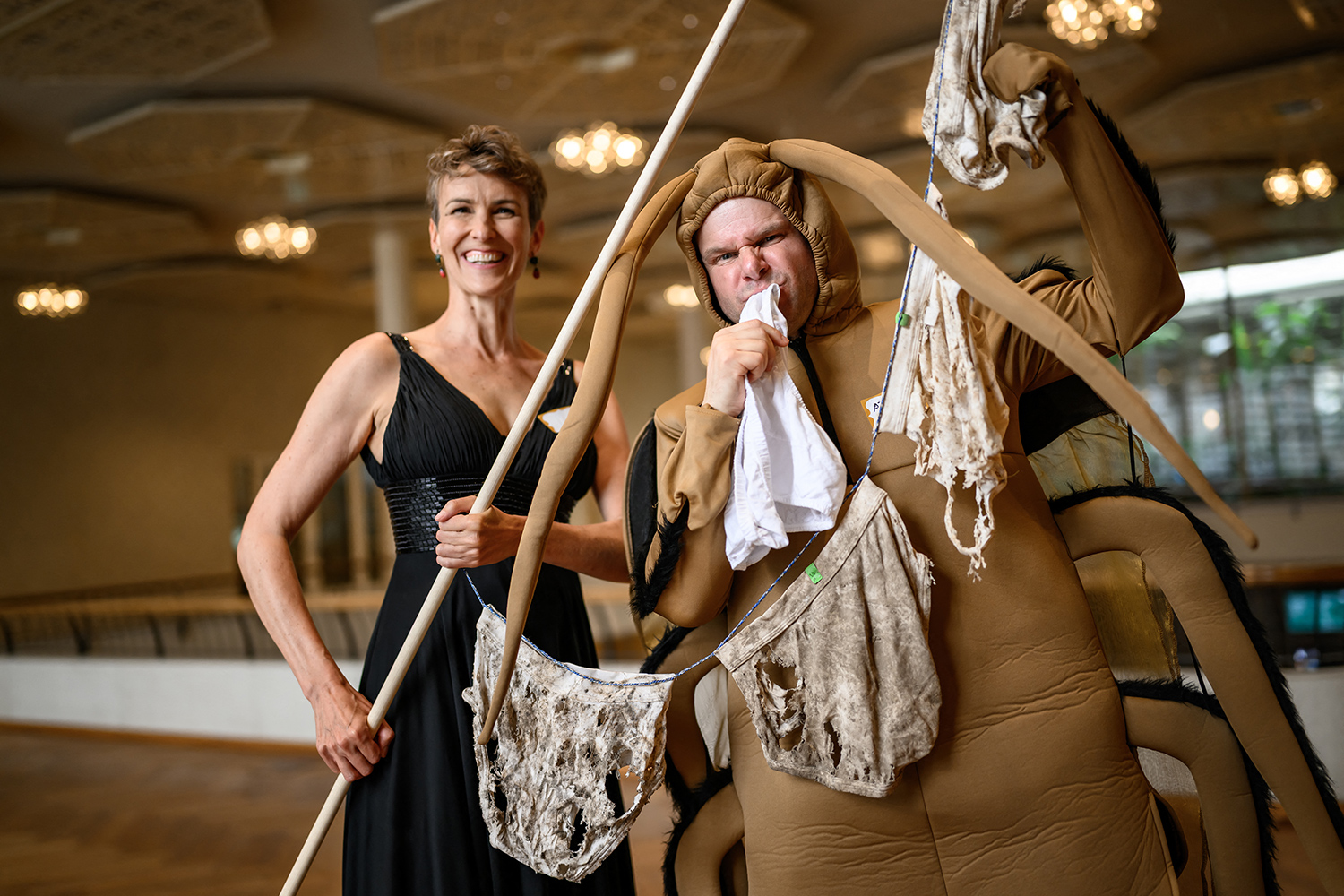 A woman in a dress and a man in a cockroach costume pose with several pairs of underwear that are attached to a line, dirty, and full of holes.