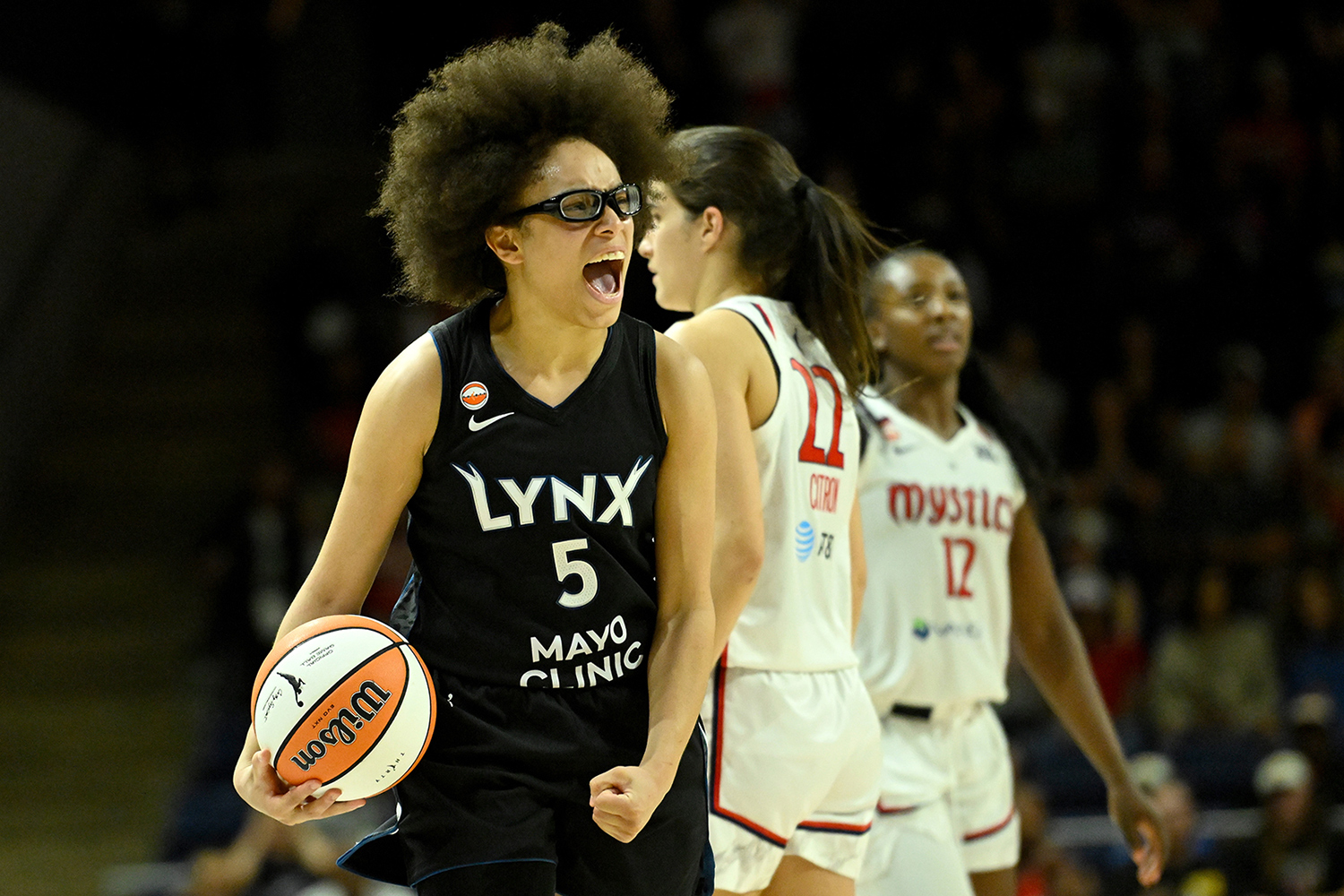 Olivia Miles makes an angry or frustrated expression while holding a basketball on a court with two players from the Washington Mystics in the background.