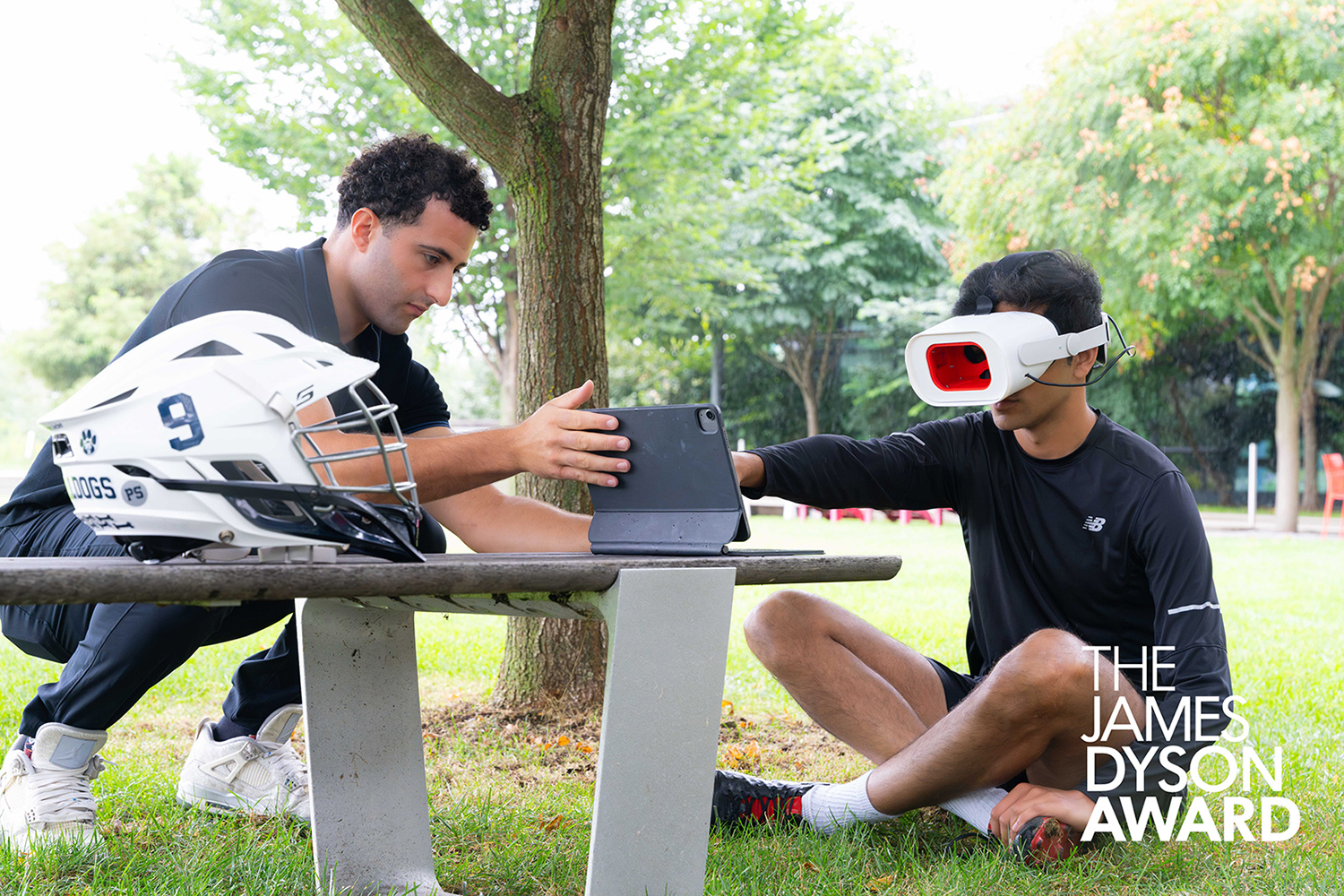 A teen sits on the grass wearing a Rumble Rims headset over his eyes in front of a table with other equipment that is being adjusted by a man.