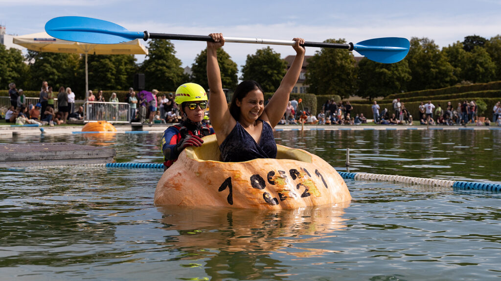 A woman raises her oar while sitting in a hollowed out giant pumpkin on a body of water as another woman holds the pumpkin from the back.
