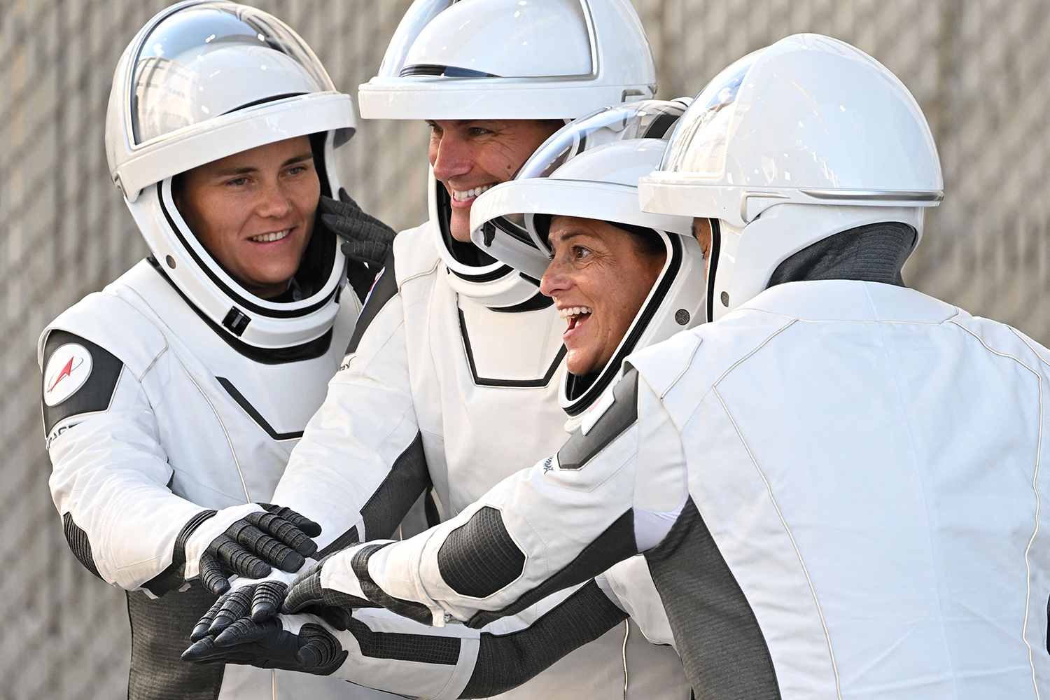 Nicole Mann and three other astronauts pose with their hands touching while wearing white astronaut uniforms.