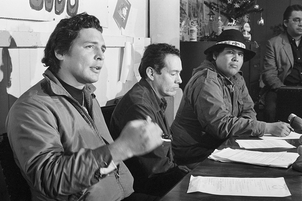 Three men sit at a table with papers in front of them. One of the men is speaking.