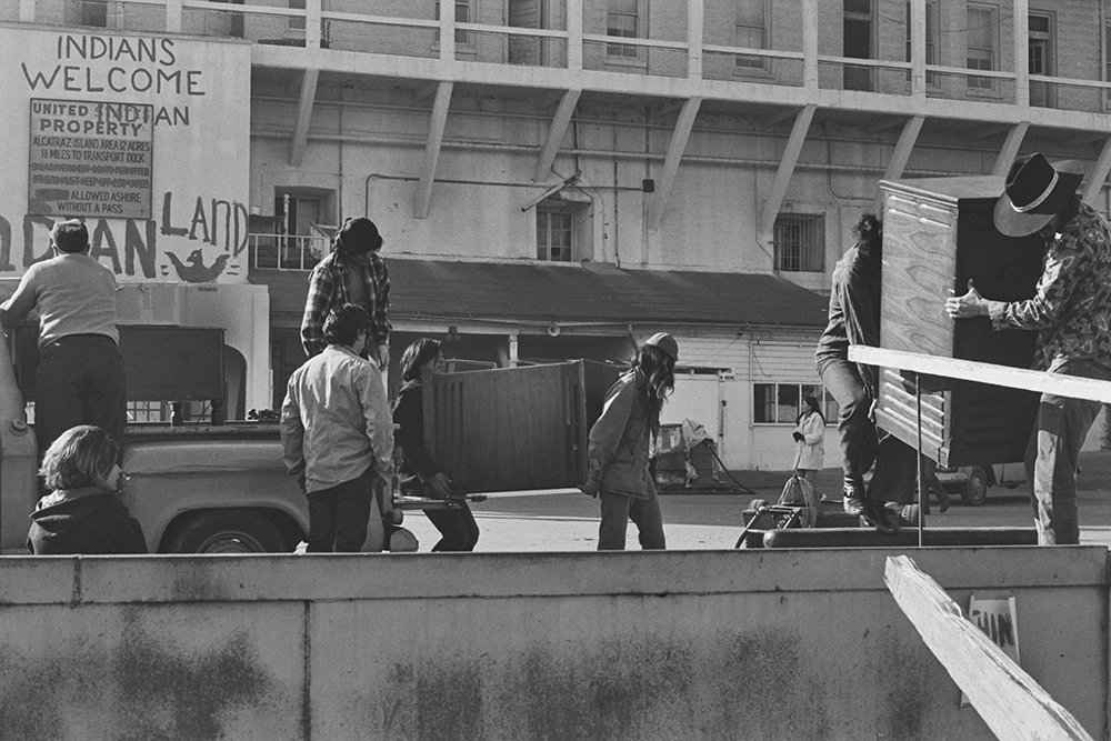 People move furniture off a pickup truck with a prison structure and a sign reading Indians Welcome and Indian Land in the background.