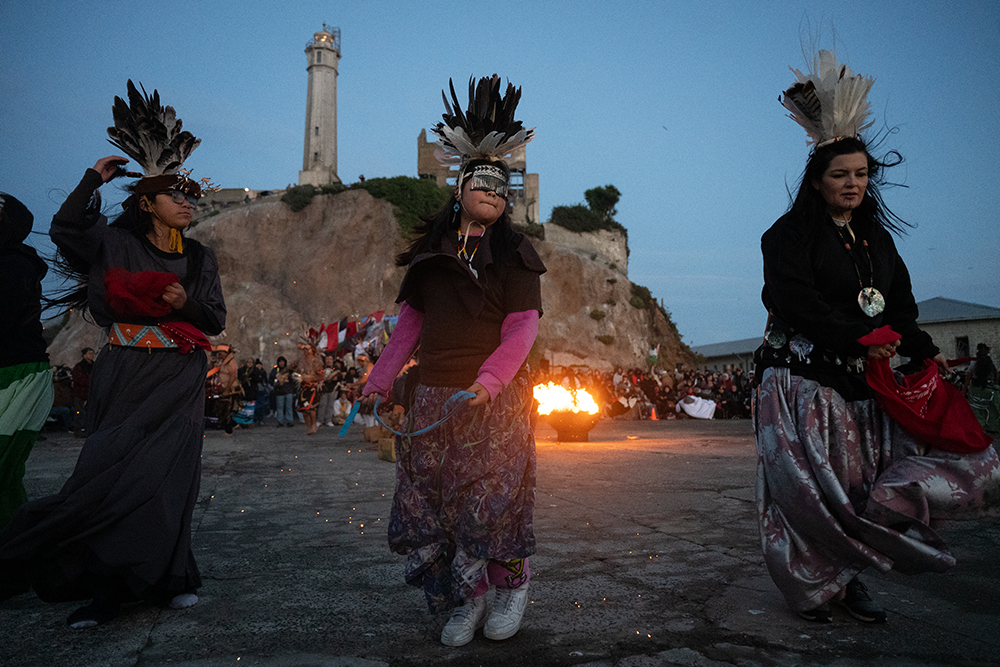 Three women wearing beads and feather headdresses dance in front of a fire and a large crowd of people with a former prison tower in the background.