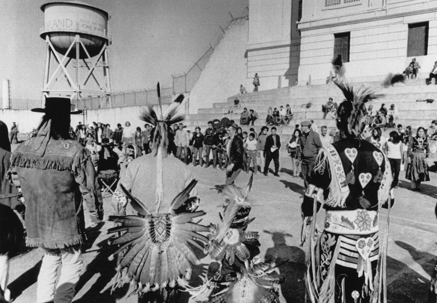 A group of Native American people, some wearing headdresses, gather outside a building on Alcatraz.
