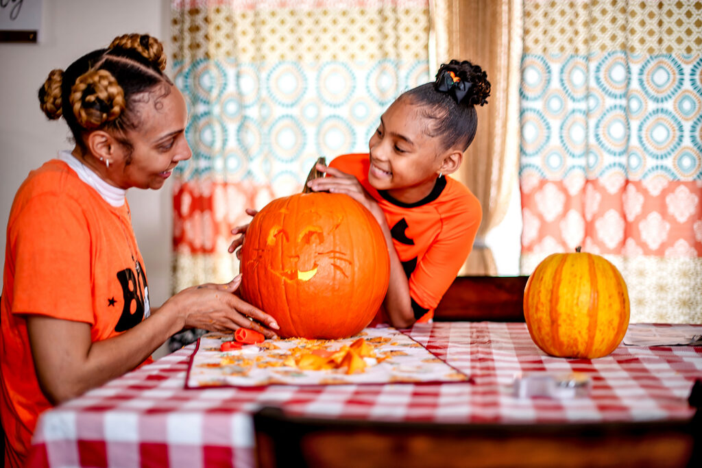 A woman and girl sit at a table holding a recently carved pumpkin and smile at each other.