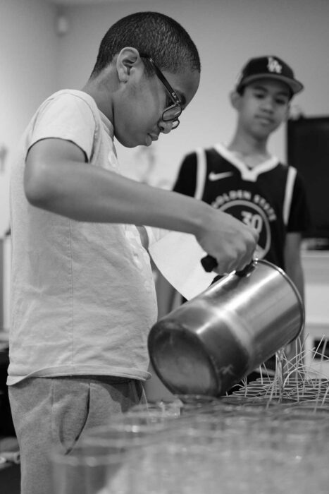 A boy pours something from a pitcher into glass containers as another boy watches.